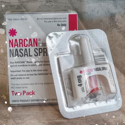 A Narcan Nasal Spray applicator in the packaging leaning on a box of Narcan Nasal Spray