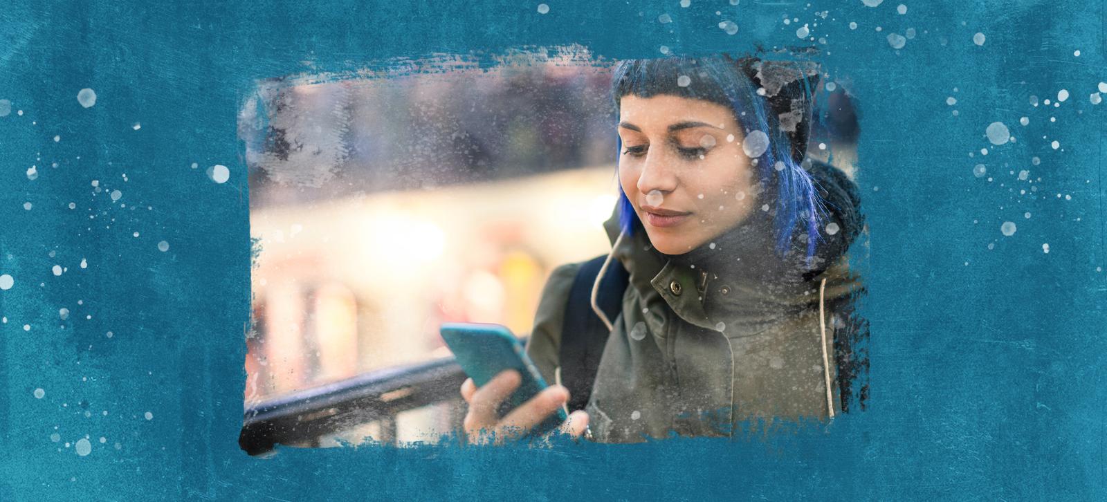 A girl is walking and looking at her phone. She has blue hair, is wearing an army green jacket, and has a backpack on.