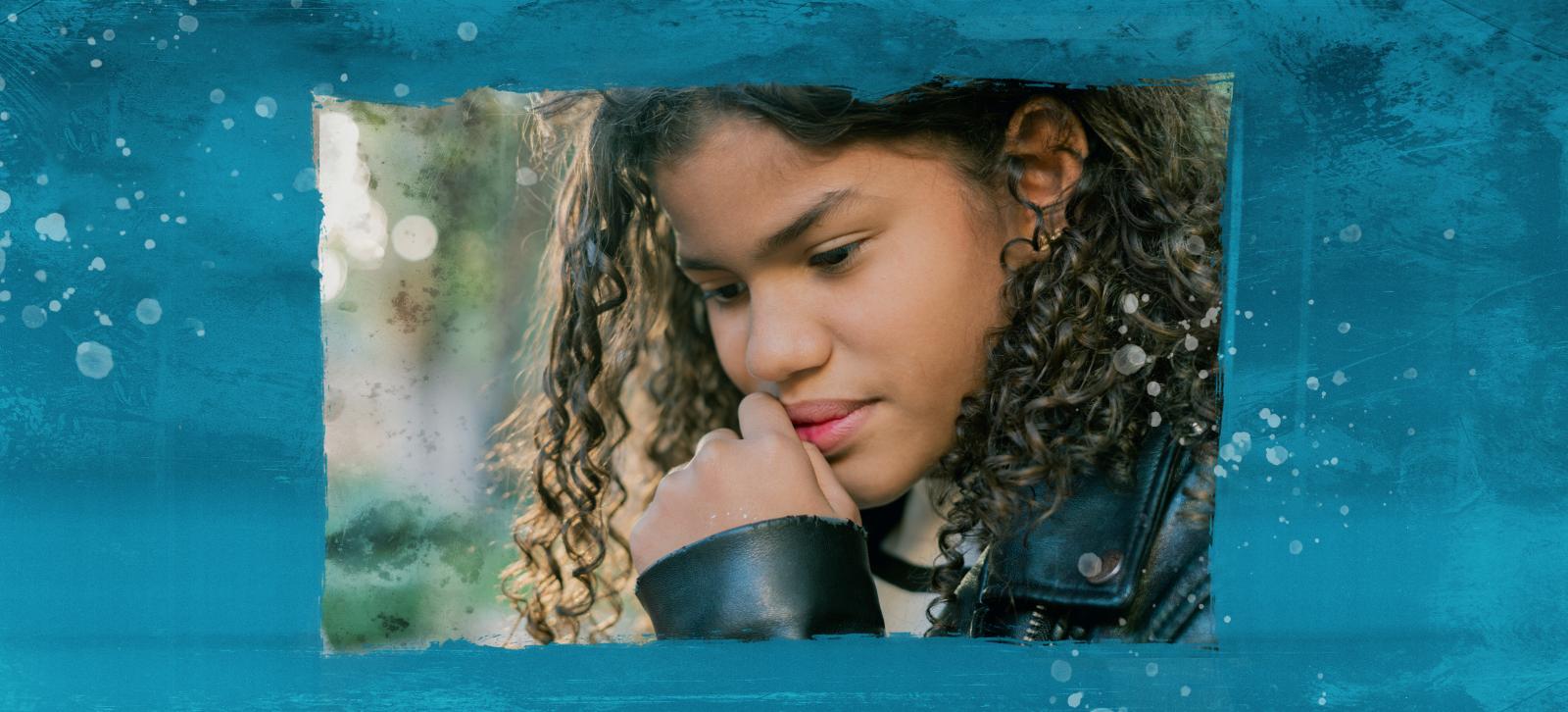 A girl with curly hair is deep in thought, looking at the ground with her hand to her mouth.