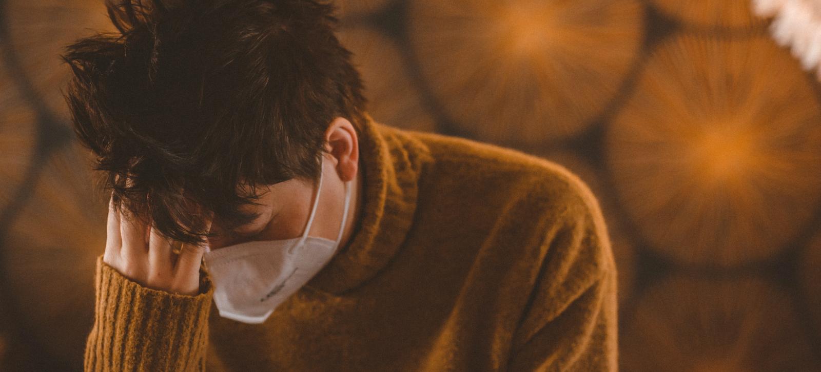 Person in a mask looking down, holding their head.