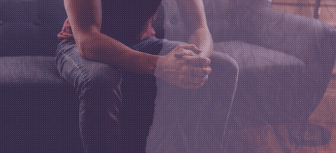  An image from the shoulders down of a person sitting on a gray couch. The image has a distressed purple tint over it with the left half of the image being darker than the right.